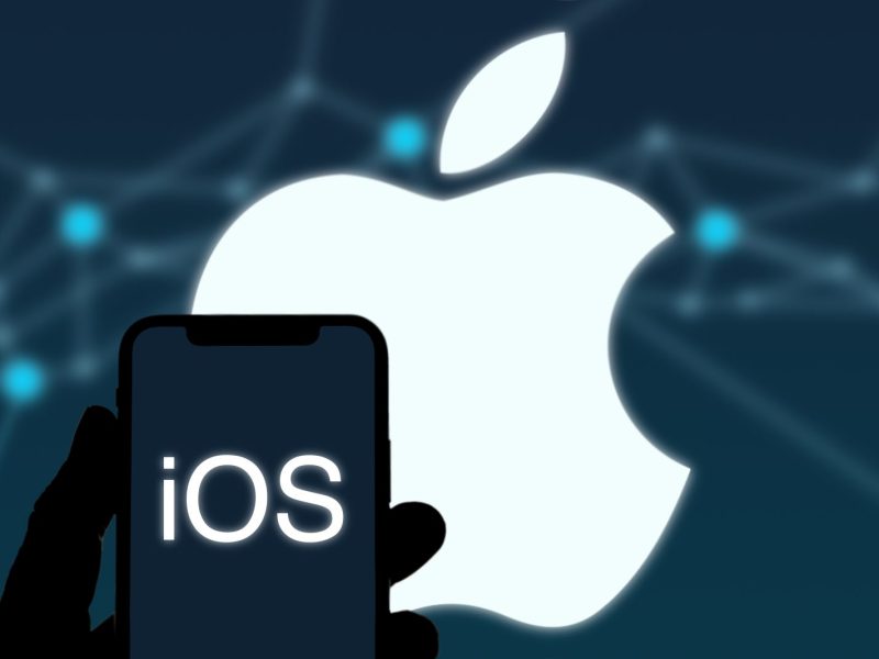 iOS-Logo auf einem Handy-Bildschirm, im Hintergrund ist das Apple-Logo zu sehen.