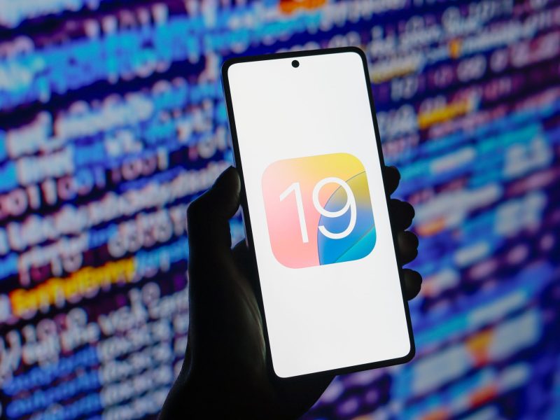 Person hält Smartphone in der Hand. Auf dem Bildschirm das Logo von iOS 19.