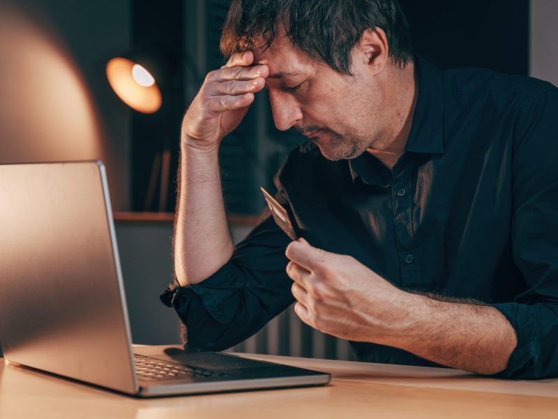 Mann hält verzweifelt die Hand an den Kopf, während er vor einem Laptop sitzt, mit einer Bankkarte in der Hand