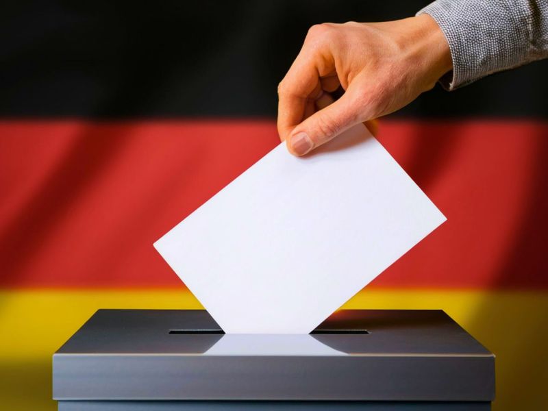 Person wirft Zettel in die Wahlurne (vor einer Deutschlandfahne)