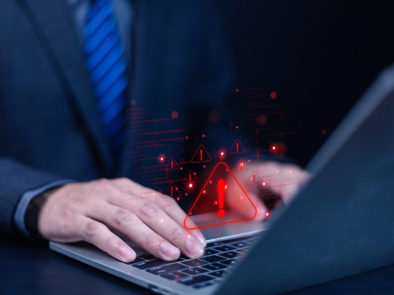 Person tippt an einem Laptop. Über dem Bild liegt ein rotes Warndreieck mit Ausrufezeichen in der Mitte.