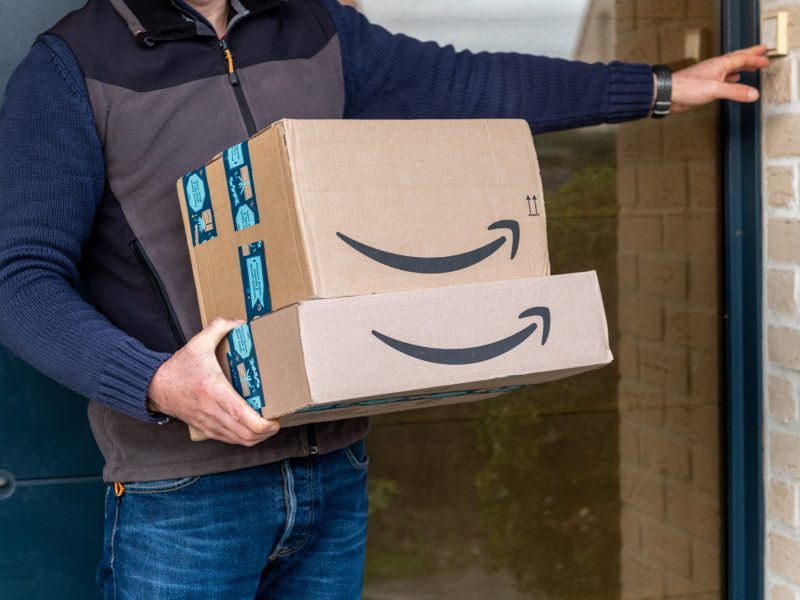 Person hält Amazon-Pakete in der Hand und klingelt an einer Tür.