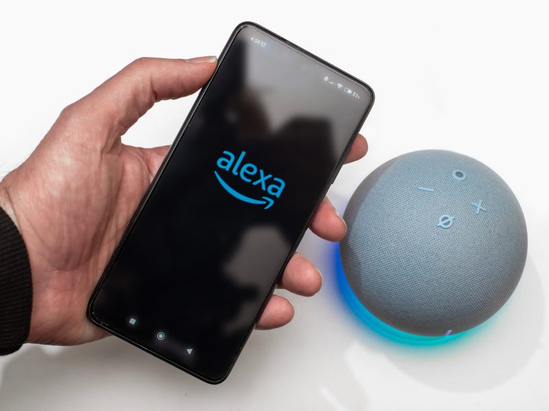 Person hält ein Handy, auf dessen Display das Alexa-Logo angezeigt wird. Daneben ist ein Amazon Echo Dot zu sehen.