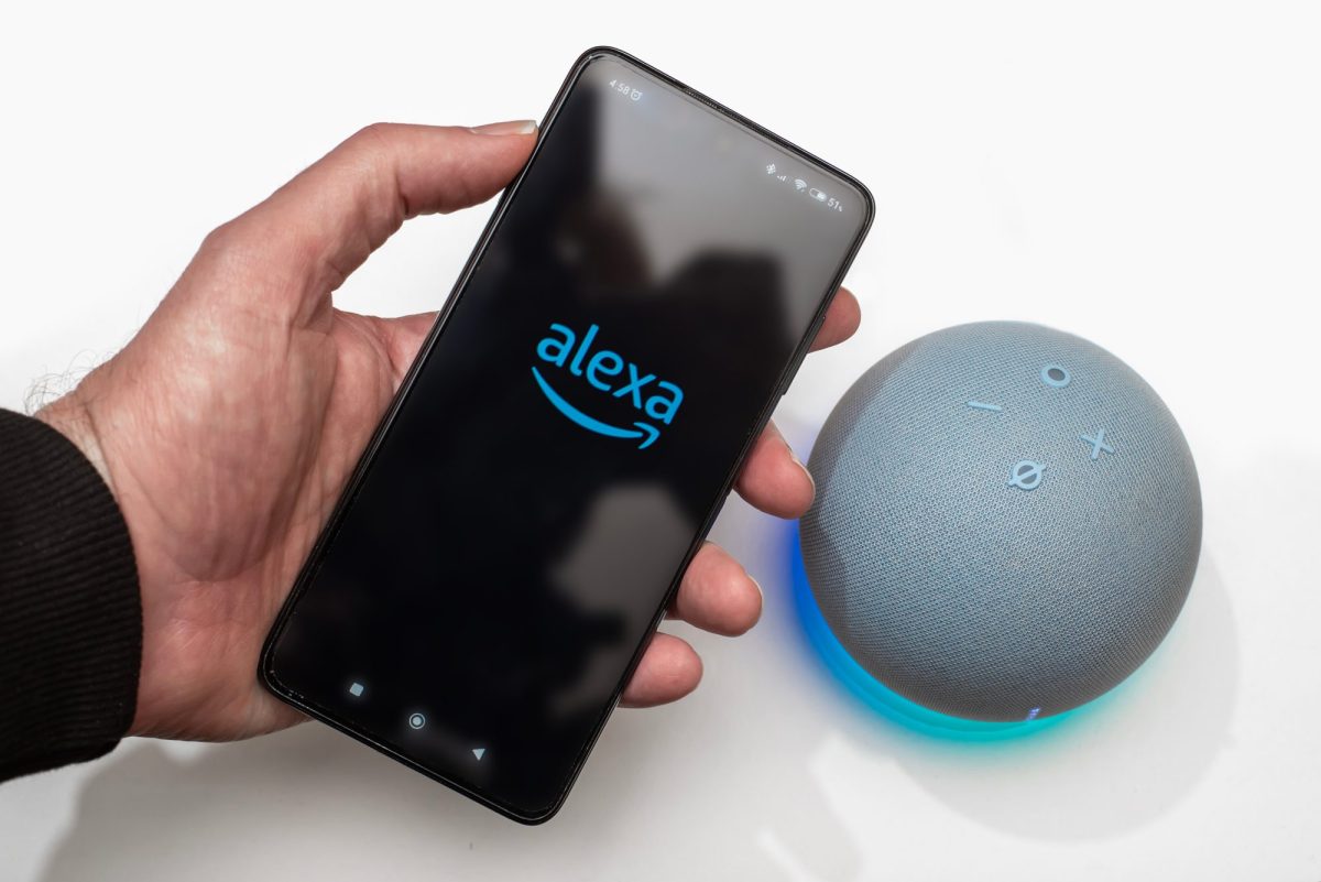 Person hält ein Handy, auf dessen Display das Alexa-Logo angezeigt wird. Daneben ist ein Amazon Echo Dot zu sehen.