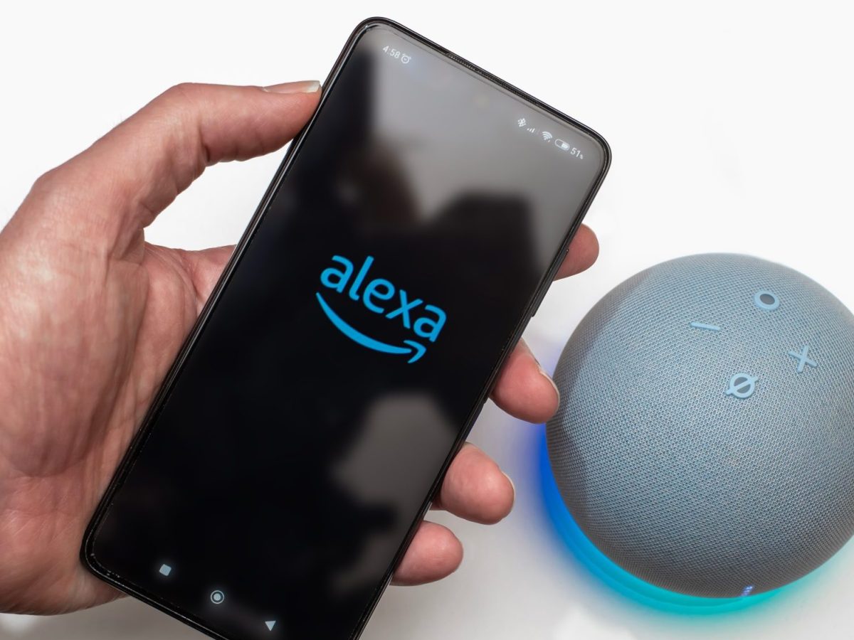 Person hält ein Handy, auf dessen Display das Alexa-Logo angezeigt wird. Daneben ist ein Amazon Echo Dot zu sehen.