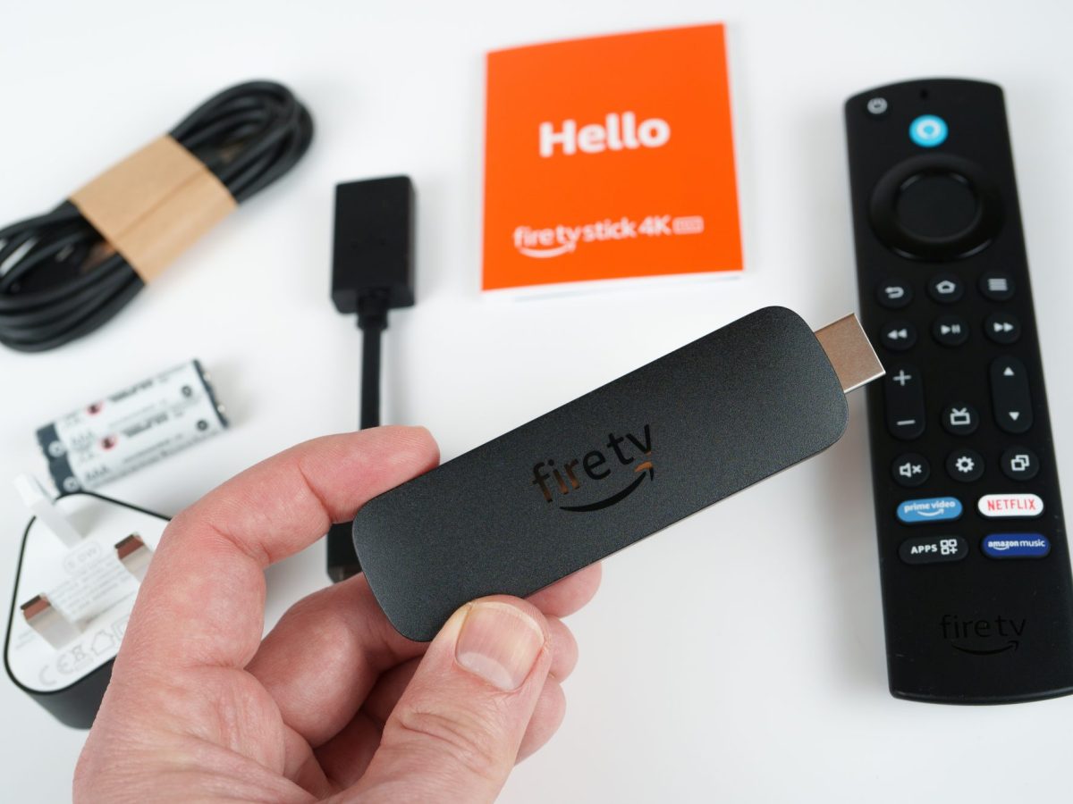 Person hält einen Amazon Fire TV-Stick. IM Hintergrund liegt das entsprechende Zubehör.