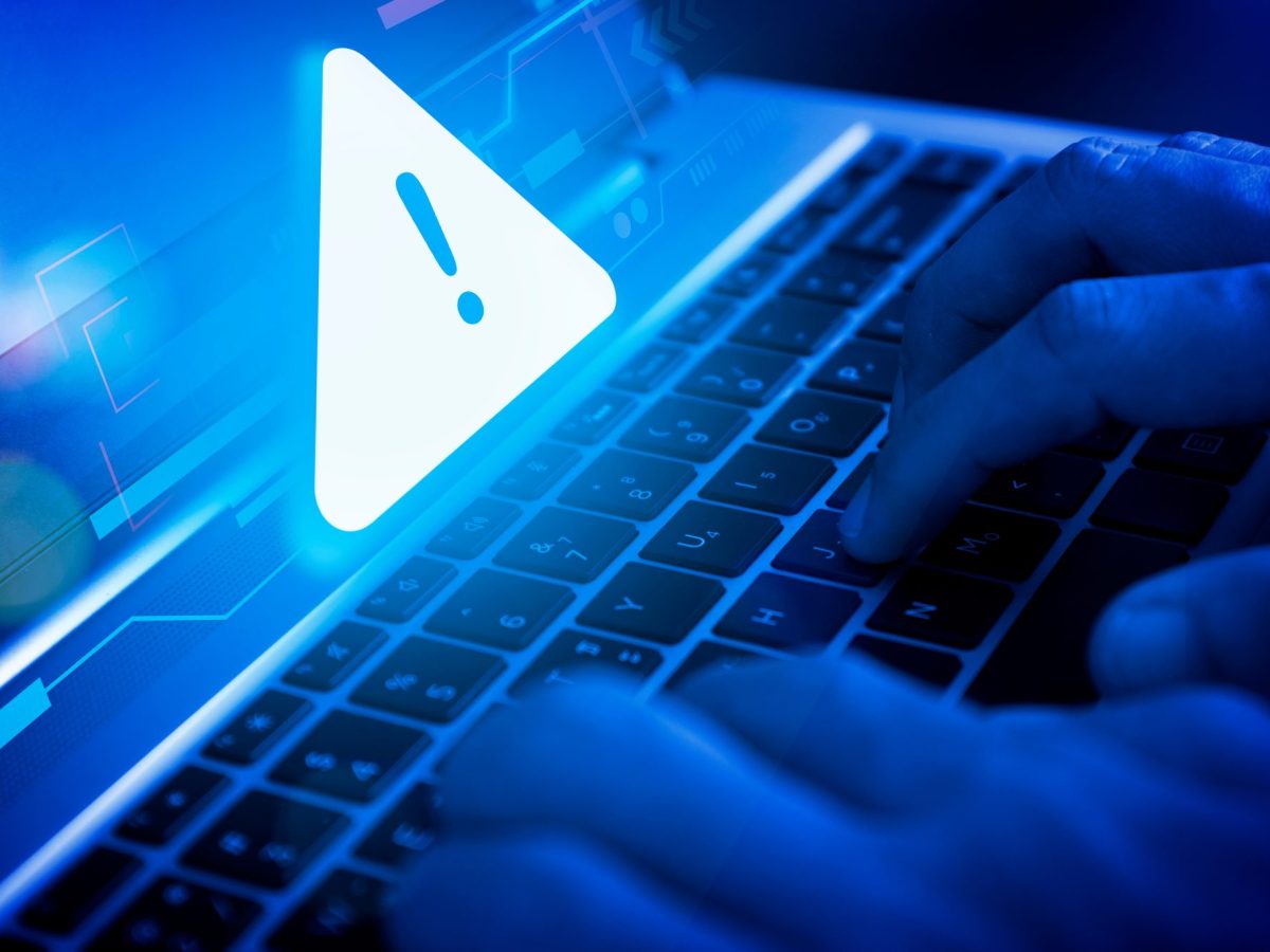 Person tippt an einem Apple-Laptop. Ãœber dem Bildschirm schwebt ein blaues Warnschild.