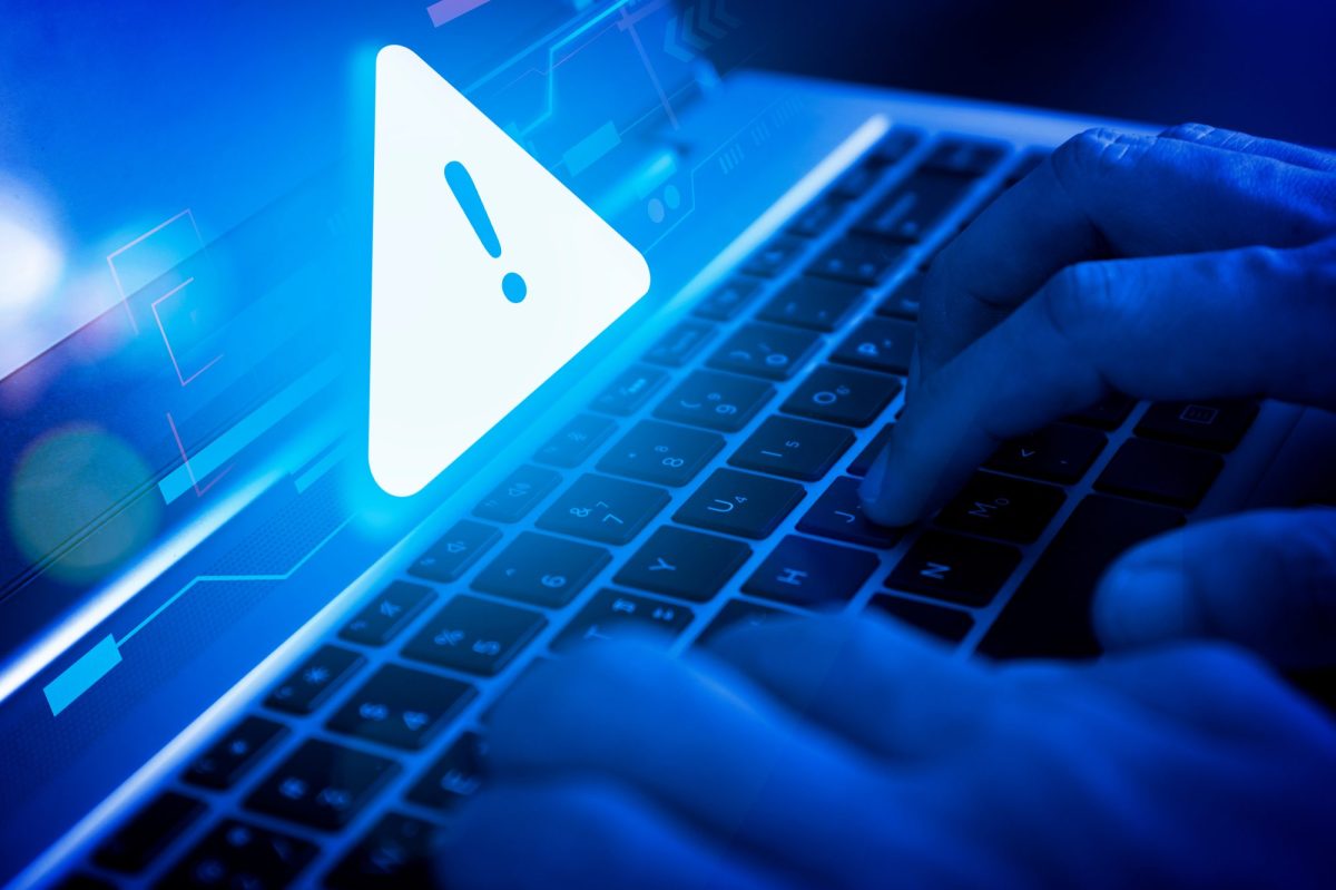 Person tippt an einem Apple-Laptop. Über dem Bildschirm schwebt ein blaues Warnschild.