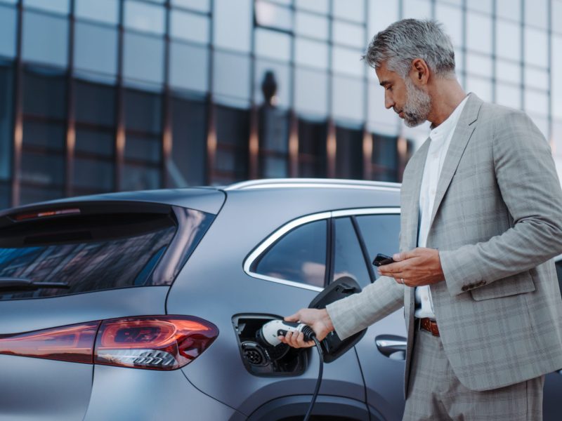 Eine Person im Anzug hält ein Handy in der Hand und lädt ein Elektroauto. Im Hintergrund ist ein modernes Gebäude mit Glasfront zu sehen.