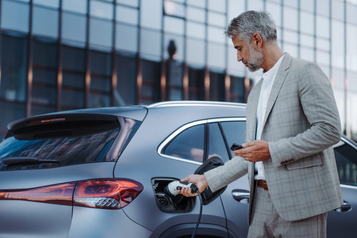 Eine Person im Anzug hält ein Handy in der Hand und lädt ein Elektroauto. Im Hintergrund ist ein modernes Gebäude mit Glasfront zu sehen.