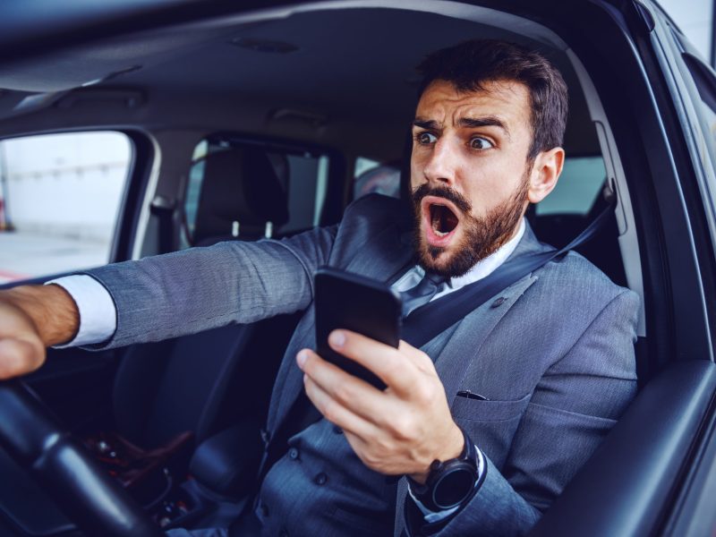 Mann hält ein Handy am Steuer und erschrickt sich.