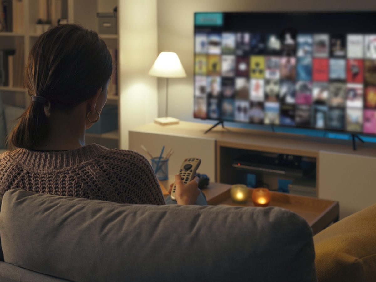 Frau sitzt auf einem Sofa und guckt auf einen eingeschalteten Fernseher.