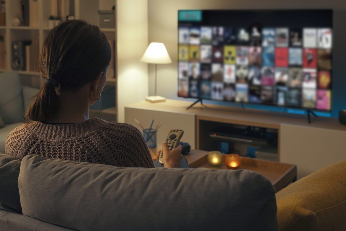 Frau sitzt auf einem Sofa und guckt auf einen eingeschalteten Fernseher.