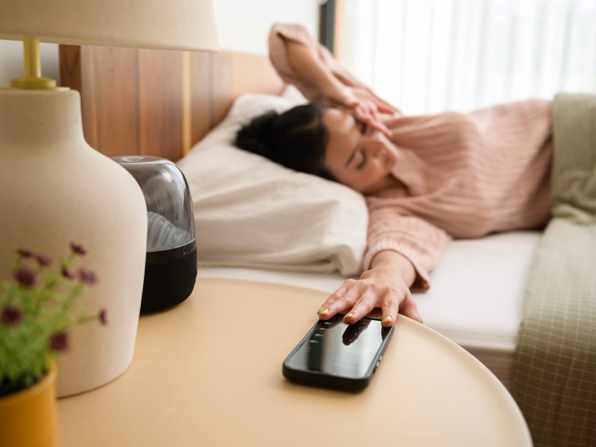 Eine Person liegt im Bett und greift zum Handy, das auf einem Tisch neben dem Bett liegt.