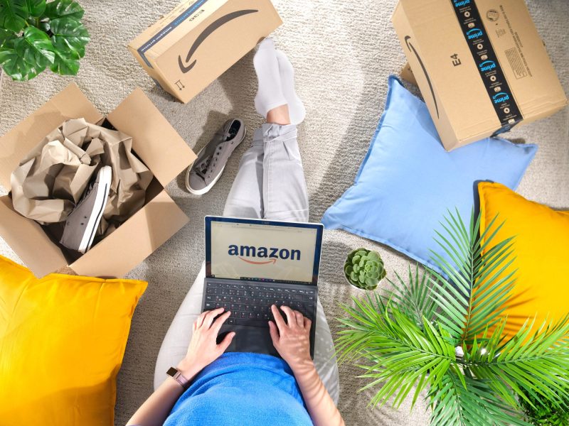 Person bedient einen Laptop. Auf dessen Display wird das Amazon-Logo angezeigt. Daneben liegen Amazon-Kartons.