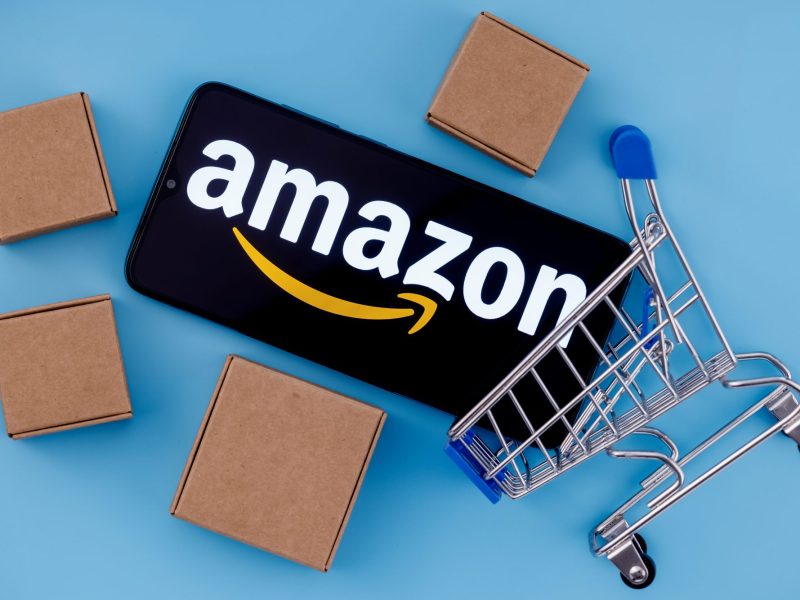 Handy mit Amazon-Logo liegt neben einem Einkaufskorb und Kartons.