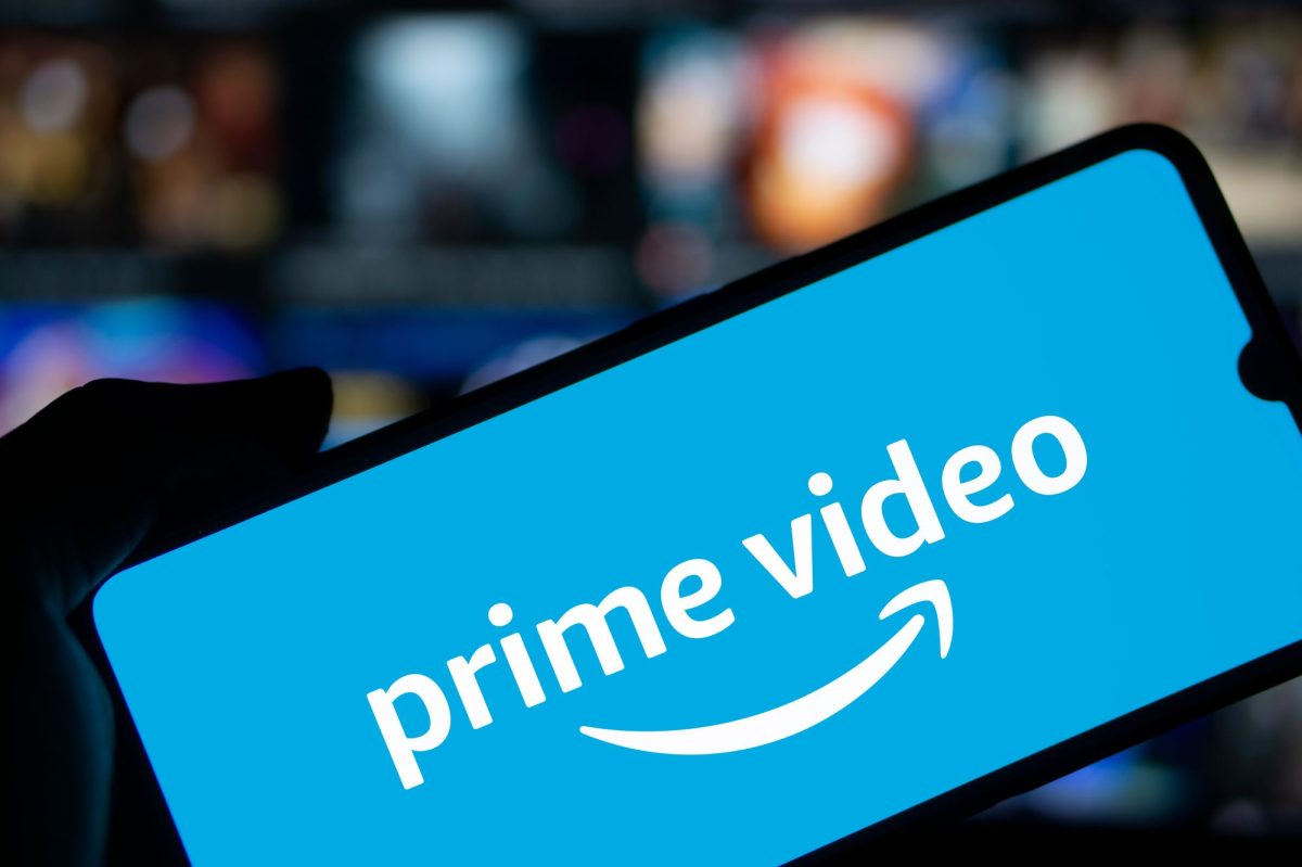 Amazon Prime Video-Logo wird auf einem Handy-Display angezeigt. Dahinter ist ein eingeschalteter Fernseher zu sehen.