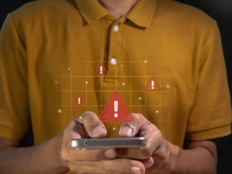 Person hält ein Handy in der Hand. Darüber ist ein rotes Ausrufezeichen zu sehen und symbolisiert Gefahr.