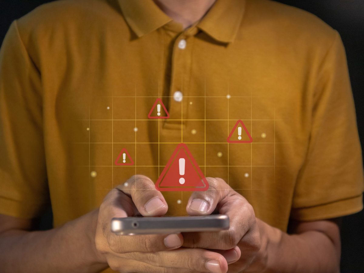 Person hält ein Handy in der Hand. Darüber ist ein rotes Ausrufezeichen zu sehen und symbolisiert Gefahr.