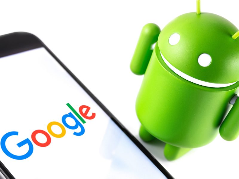 Android-Maskottchen steht neben einem Handy, auf dessen Bildschirm das Google-Logo zu sehen ist.
