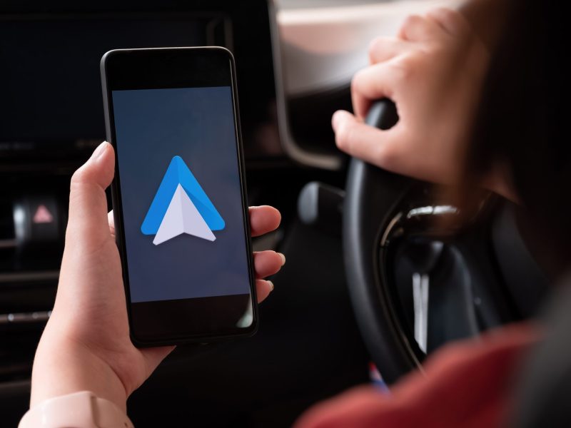 Android Auto wird auf dem Display eines Handys angezeigt. Im Hintergrund ist eine Person am Steuer eines Fahrzeugs zu sehen.