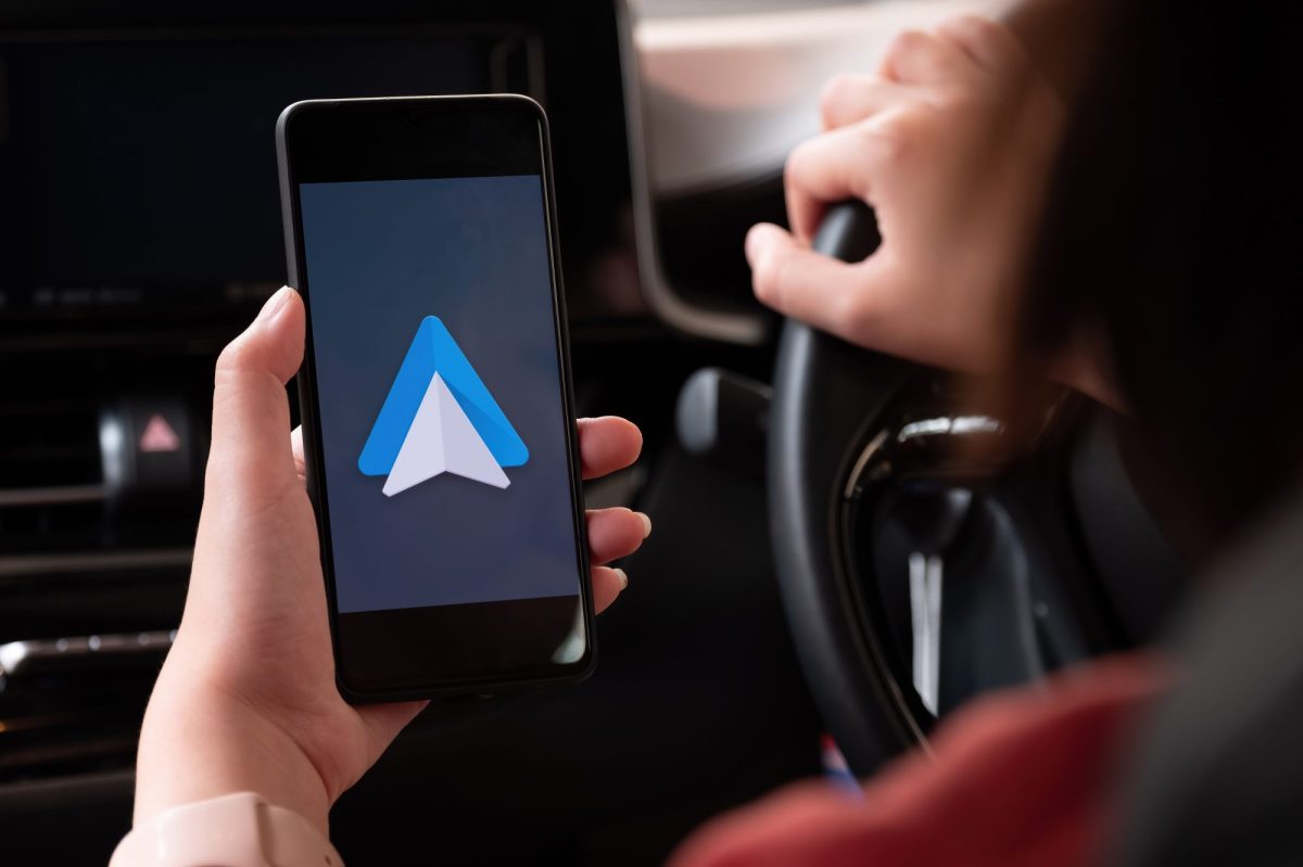 Android Auto wird auf dem Display eines Handys angezeigt. Im Hintergrund ist eine Person am Steuer eines Fahrzeugs zu sehen.