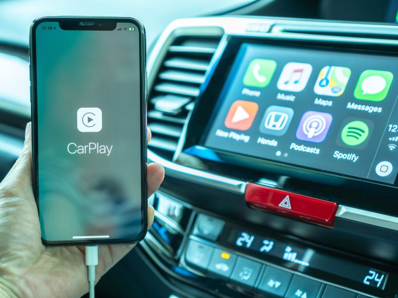 Apple CarPlay wird auf einem Handy angezeigt. Im Hintergrund ist ein Auto-Display mit verschiedenen Apps zu sehen.
