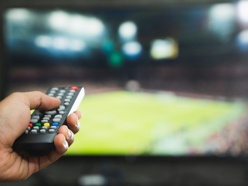 Person richtet die Fernbedienung auf einen Fernseher. Auf dem Bildschirm wird ein Fußballfeld angezeigt.
