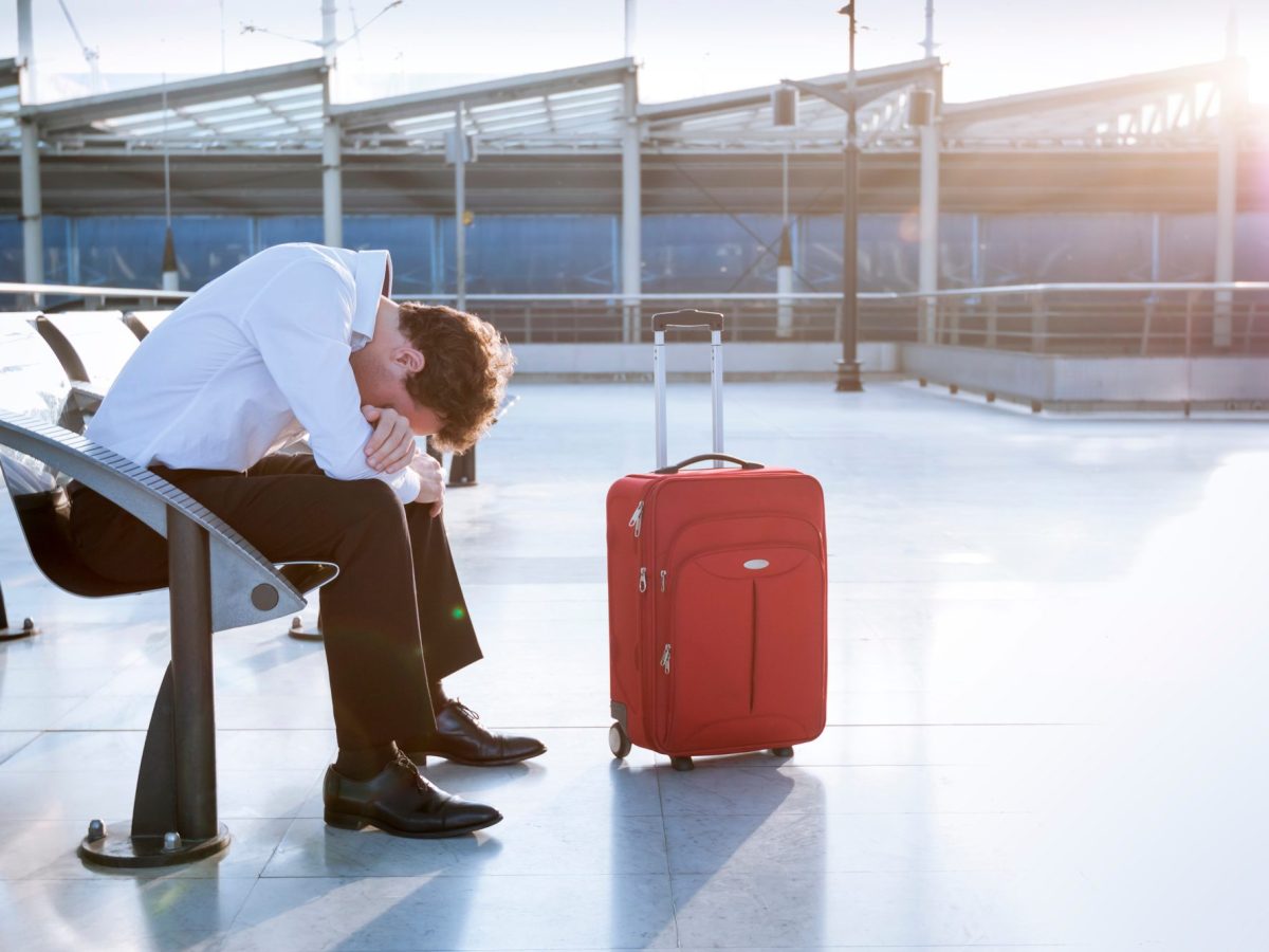 Mann sitzt in verzweifelter Haltung auf einem Flughafen. Neben ihm steht ein Koffer.