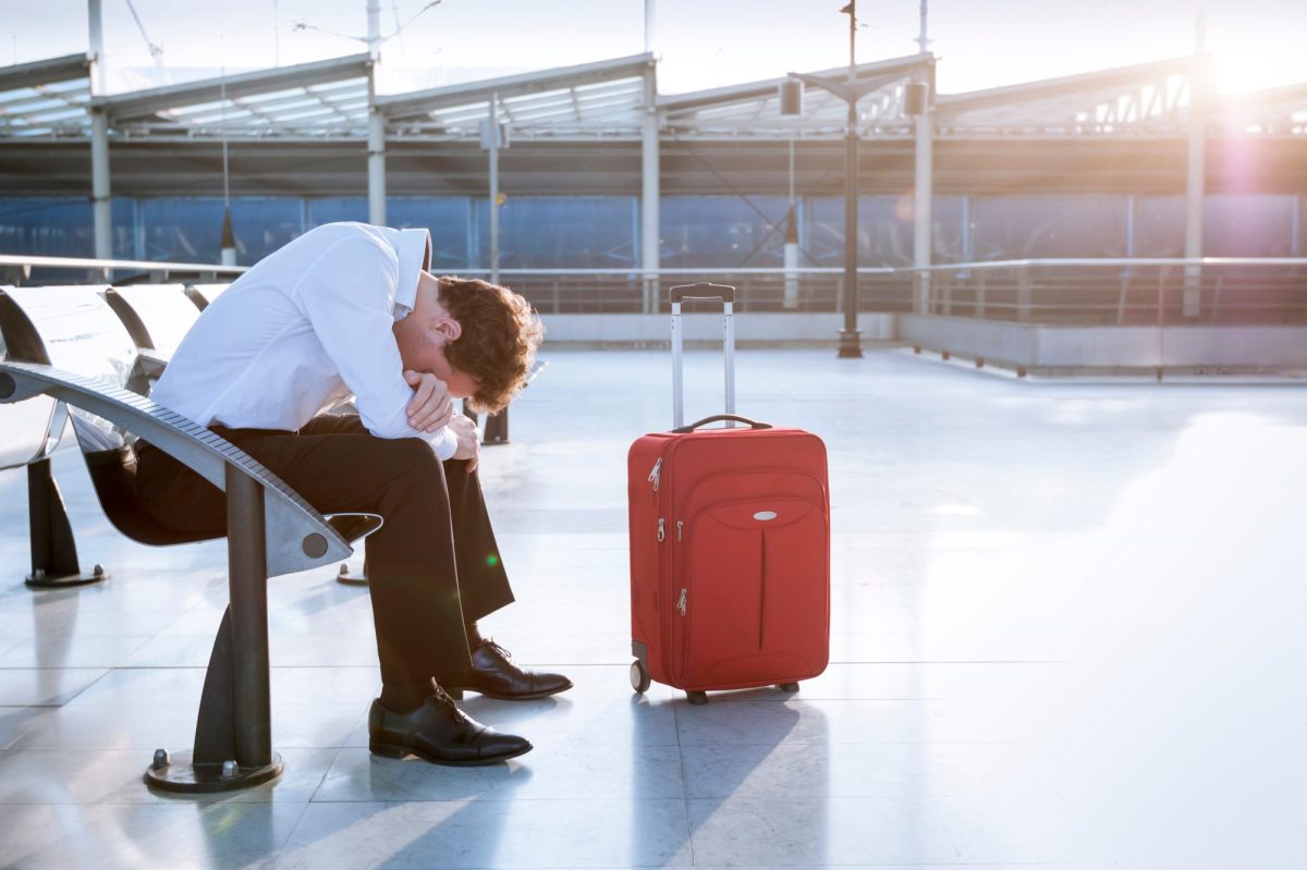 Mann sitzt in verzweifelter Haltung auf einem Flughafen. Neben ihm steht ein Koffer.