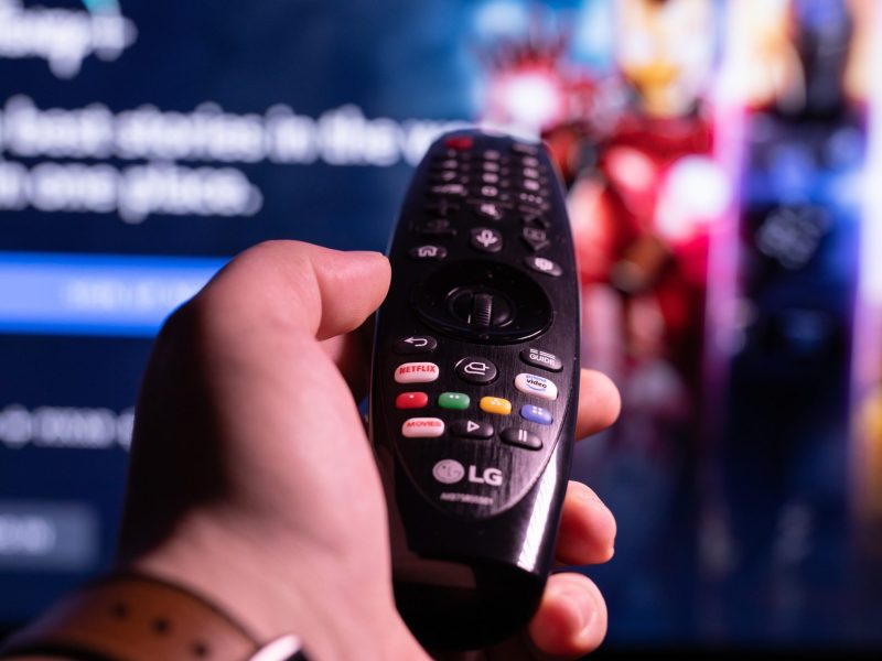 Person hält eine Fernbedienung in der Hand. Im Hintergrund ist ein eingeschalteter Fernseher zu sehen.