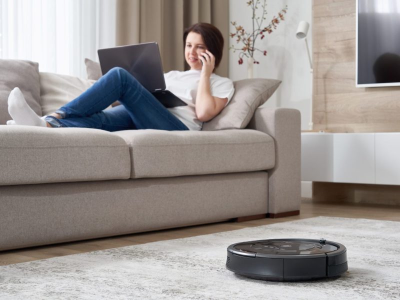 Person sitzt auf dem Sofa, während ein Saugroboter über den Boden fährt.