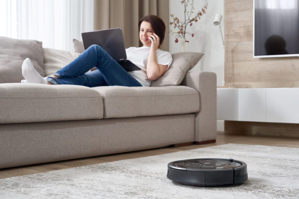 Person sitzt auf dem Sofa, während ein Saugroboter über den Boden fährt.
