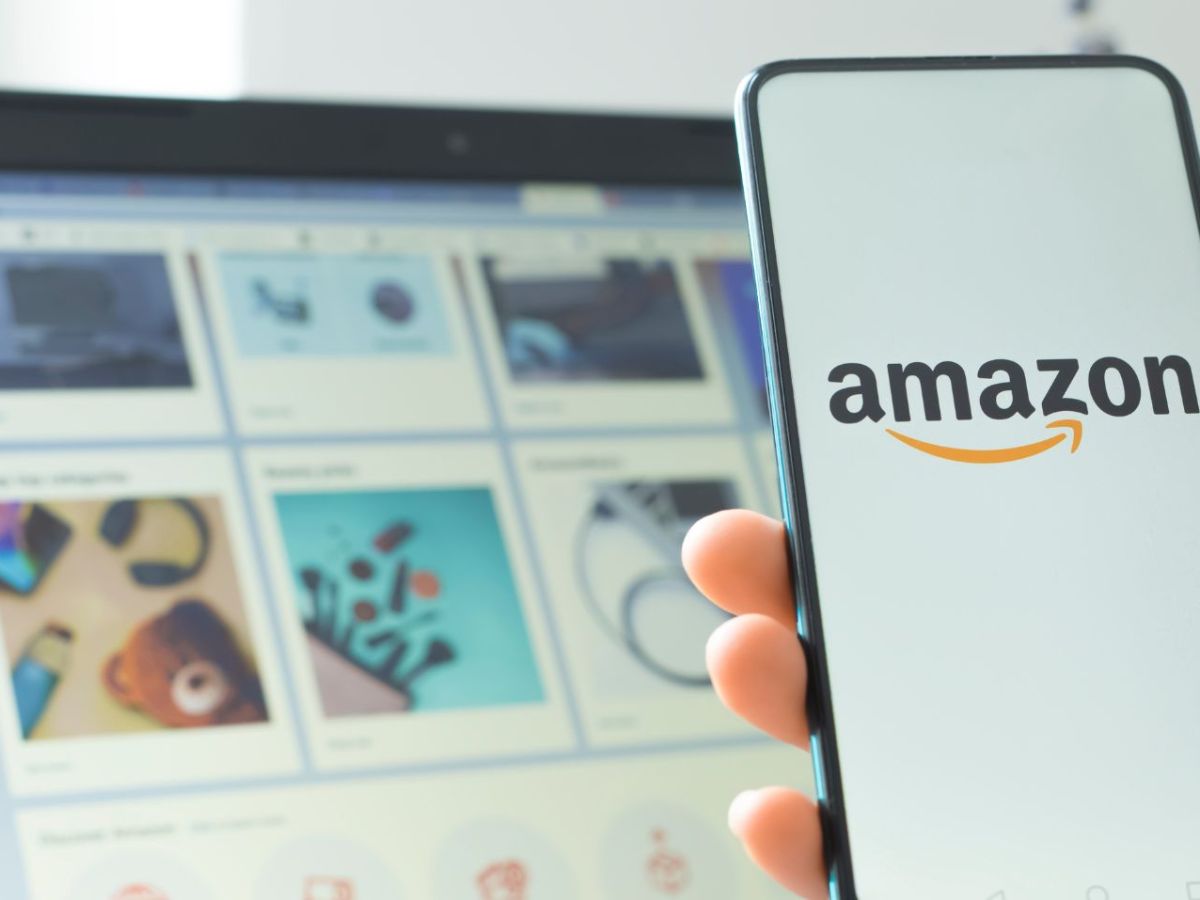 Eine Person hält ein Handy in der Hand, auf dem das Amazon-Logo angeziegt wird. Im Hintergrund ist ein Laptop zu sehen, auf dem der Amazon-Shop geöffnet ist.