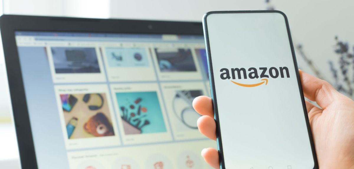 Eine Person hält ein Handy in der Hand, auf dem das Amazon-Logo angeziegt wird. Im Hintergrund ist ein Laptop zu sehen, auf dem der Amazon-Shop geöffnet ist.