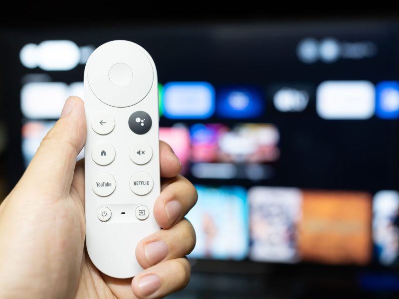 Person hält eine Google TV-Fernbedienung vor einen eingeschalteten Fernseher.