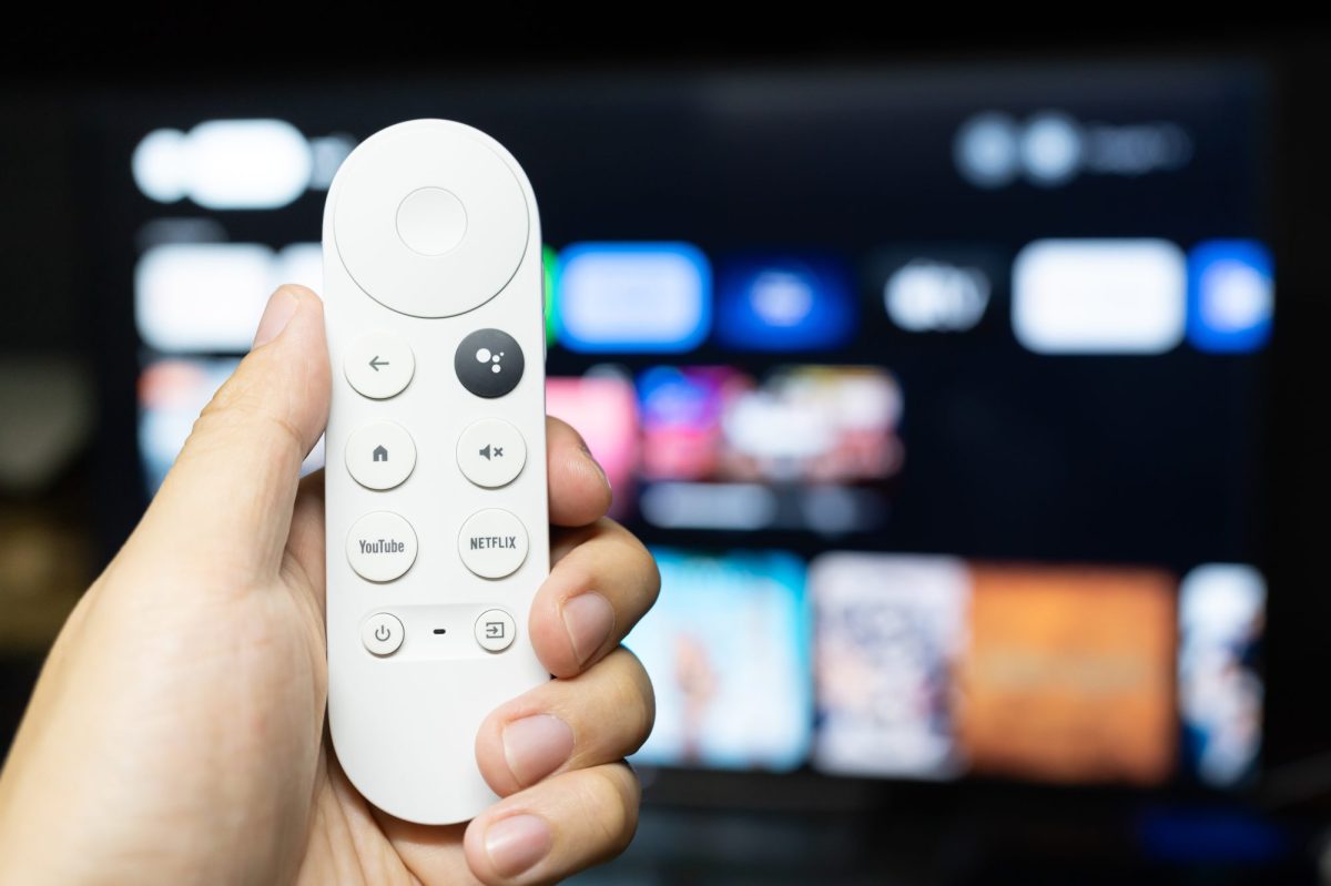 Person hält eine Google TV-Fernbedienung vor einen eingeschalteten Fernseher.