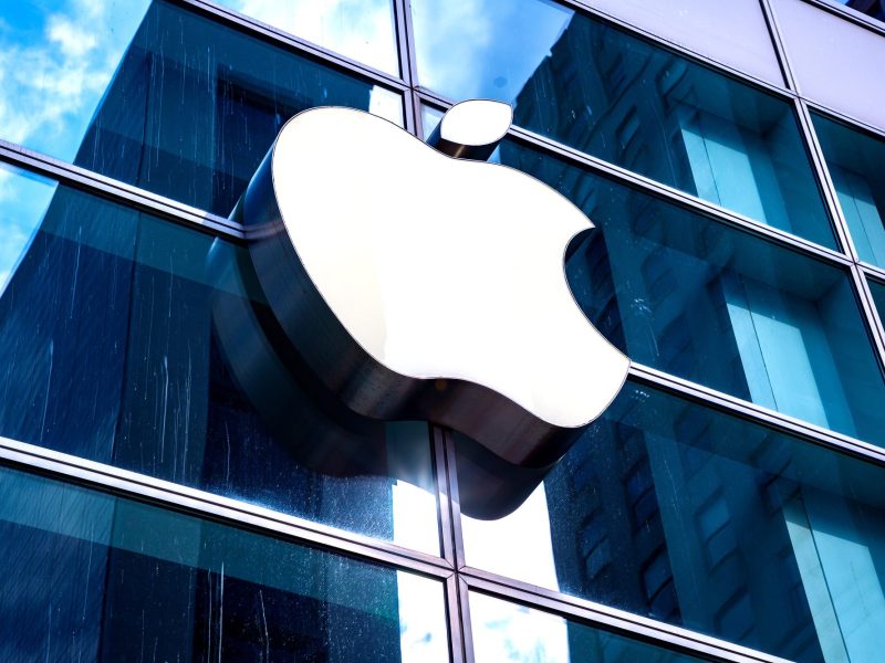 Apple-Logo an einer Glasfassade, die das Blau des Himmels reflektiert.