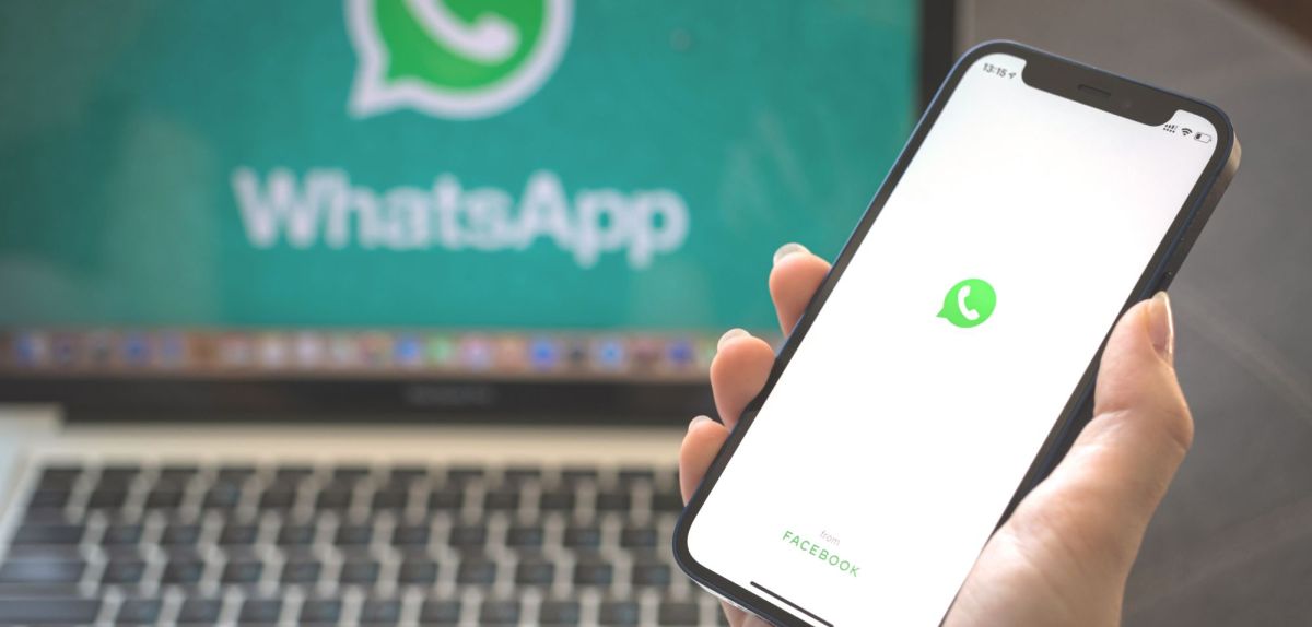 Hand hält Smartphone mit Whatsapp-Symbol vor Laptop mit demselben Symbolen.