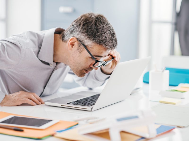 Mann mit Brille guckt intensiv auf einen Laptop, der auf einem Schreibtisch mit mehreren Dokumenten liegt.