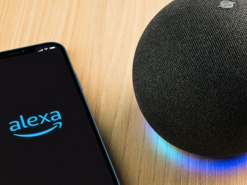 Amazon Alexa wird auf einem Handy-Display angezeigt. Daneben steht ein Amazon Echo-Gerät.