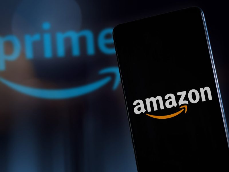 Auf einem Handy-Display ist das Amazon-Logo zu sehen. Im Hintergrund steht das Prime-Logo.