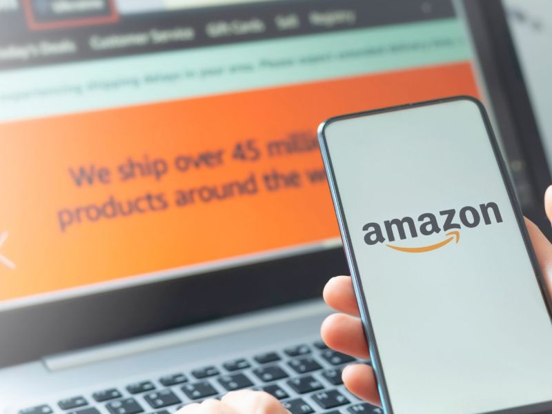 Auf einem Laptop ist der Amazon Online-Shop geöffnet. Im Vordergund hält eine Person ein Handy in der Hand, auf dem das Amazon-Logo angezeigt wird.