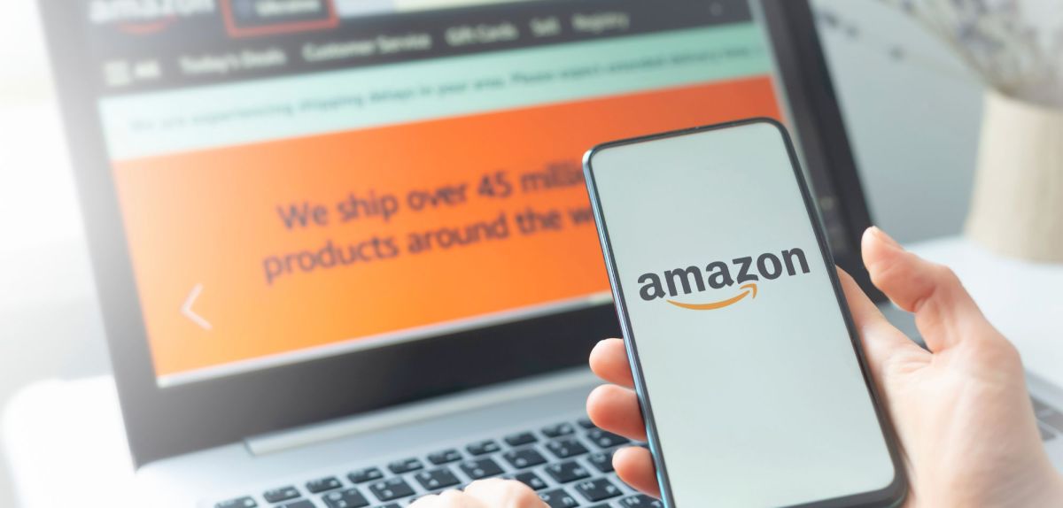 Auf einem Laptop ist der Amazon Online-Shop geöffnet. Im Vordergund hält eine Person ein Handy in der Hand, auf dem das Amazon-Logo angezeigt wird.