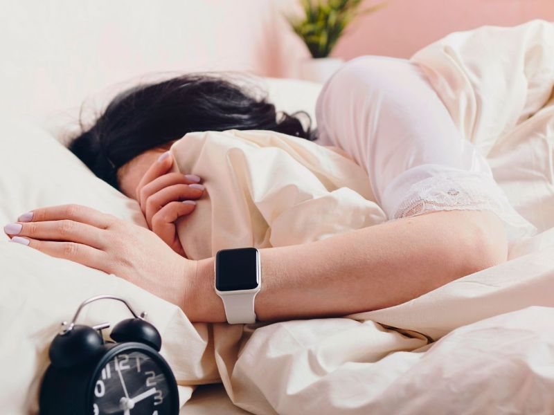 Eine Person liegt im Bett und trägt eine Apple Watch. Daneben steht ein Wecker.