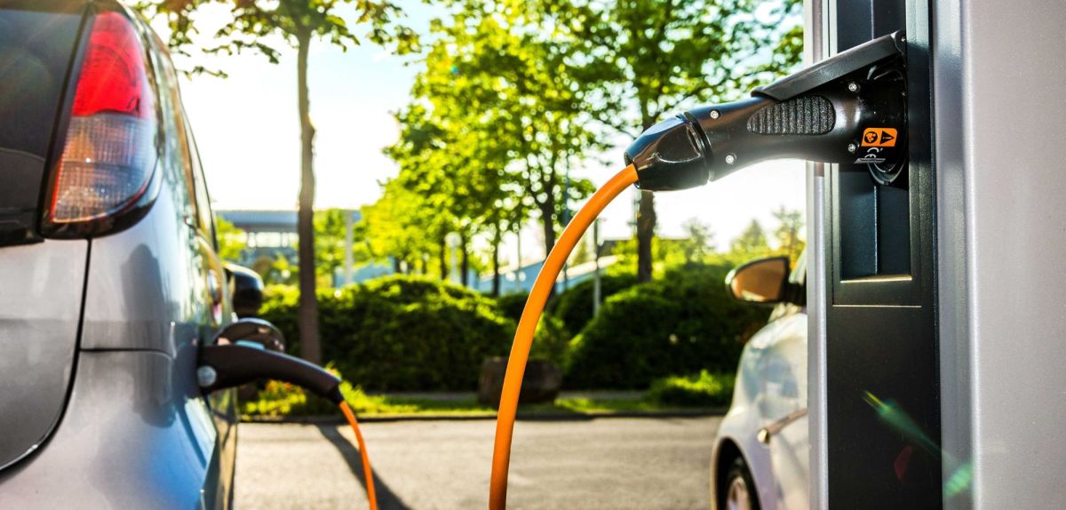 Elektroauto wird an einer Ladestation geladen. Im Hintergrund sind Bäume und eine Straße zu sehen.