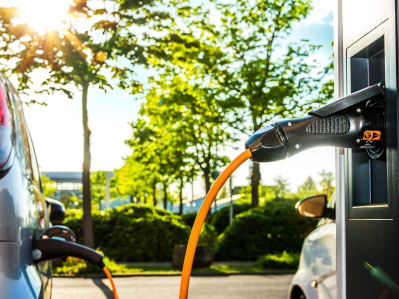 Elektroauto wird an einer Ladestation geladen. Im Hintergrund sind Bäume und eine Straße zu sehen.