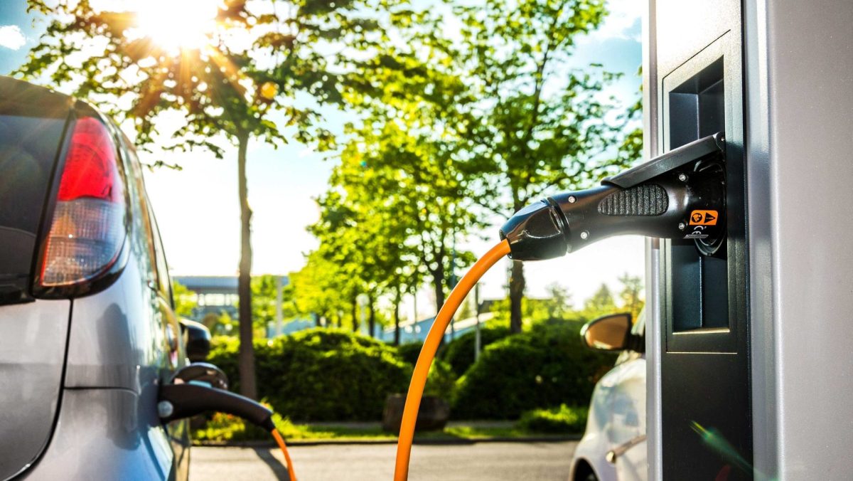 Elektroauto wird an einer Ladestation geladen. Im Hintergrund sind Bäume und eine Straße zu sehen.
