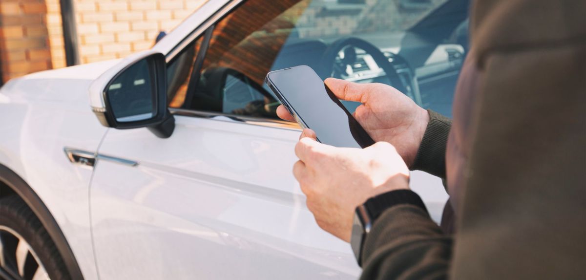 Person steht mit Smartphone in der Hand vor einem Auto.