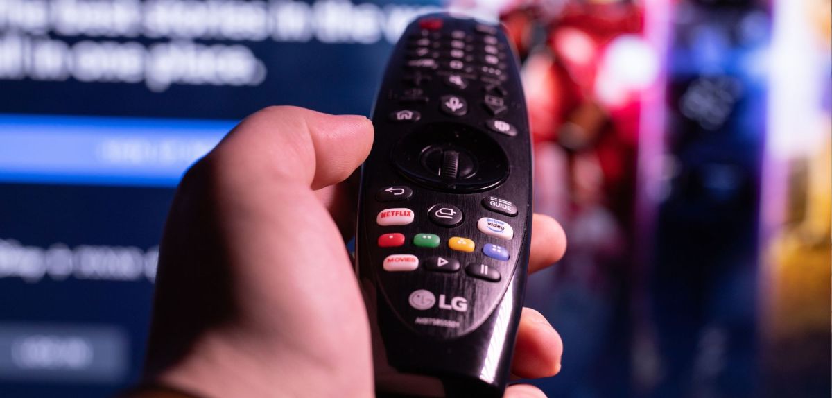 Person hält eine Fernbedienung in der Hand, die in Richtung eines Fernsehers zeigt.